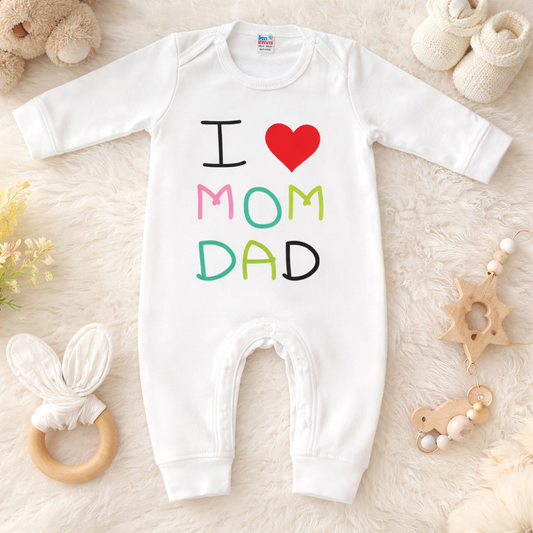 White baby onesie with 'I ❤️ MOM DAD' text on a soft surface with toys around