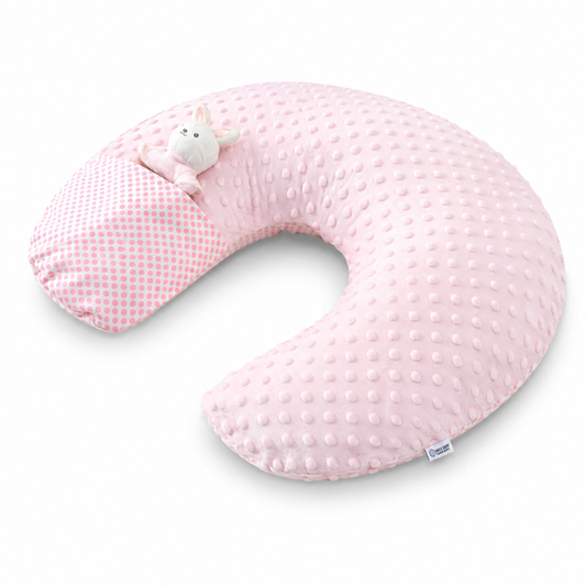 A U-shaped baby nursing pillow in light pink, featuring a textured dotty fabric on the main body and a pink-and-white gingham pattern on the side. A small plush white bunny toy is tucked into the pillow, and a small fabric brand tag is visible on the lower right edge.